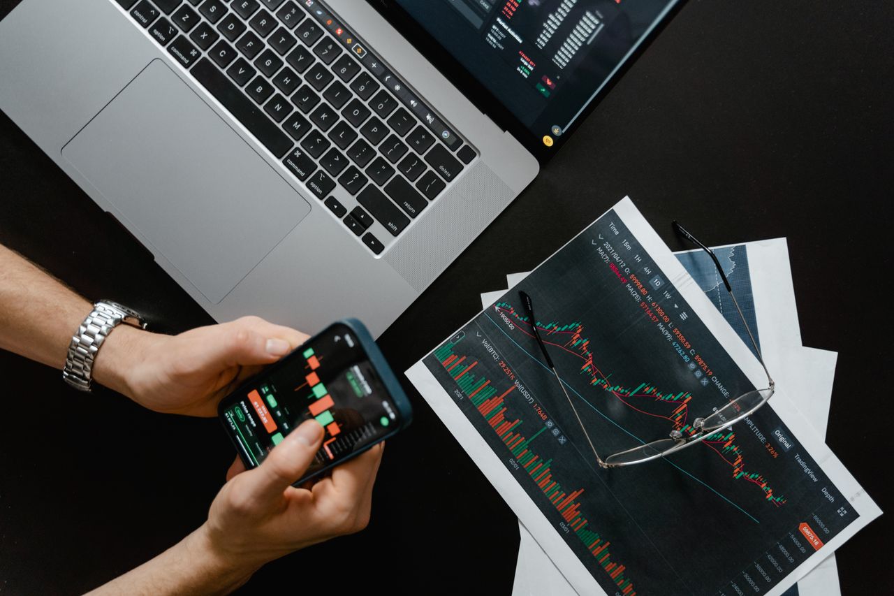 contact-img Hands holding a smartphone next to a laptop displaying stock charts, illustrating digital trading.